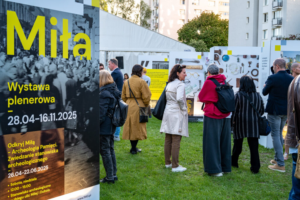 Grupa gości na wystawie "Miła", na pierwszym planie widoczna jest plansza z napisem "Miła. Wystawa plenerowa, 28.04-16.11.2025. Odkryj Miłą - Archeologia Pamięci. Zwiedzanie stanowiska archeologicznego. 26.04-22.06.2025. Sobota, niedziela, 12:00-15:00. Stanowisko archeologiczne u zbiegu ulic Miłej i Dubois"