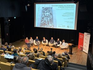 Wypełniona sala podczas wydarzenia "Zrozumieć pamięć: jak rozmawiać o trudnej przeszłości?". Na scenie widoczny ekran z banerem wydarzenia, a tuż pod nim uczestnicy debaty. Po prawej stronie od uczestników widoczne banery MGW oraz Instytutu Pileckiego.